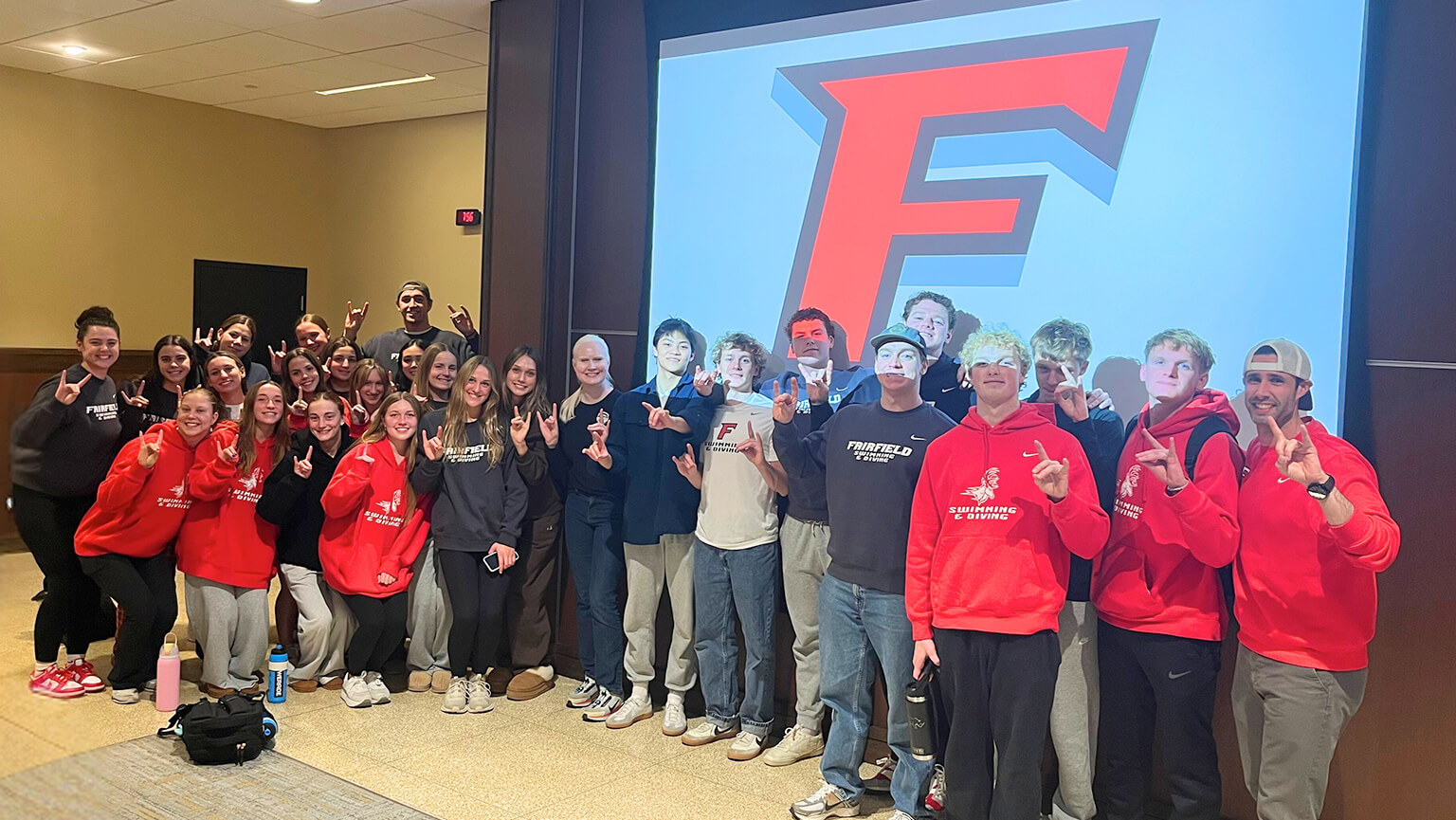 A group of students wearing red shirts stands in front of a large screen featuring the F logo, posing for a photo.