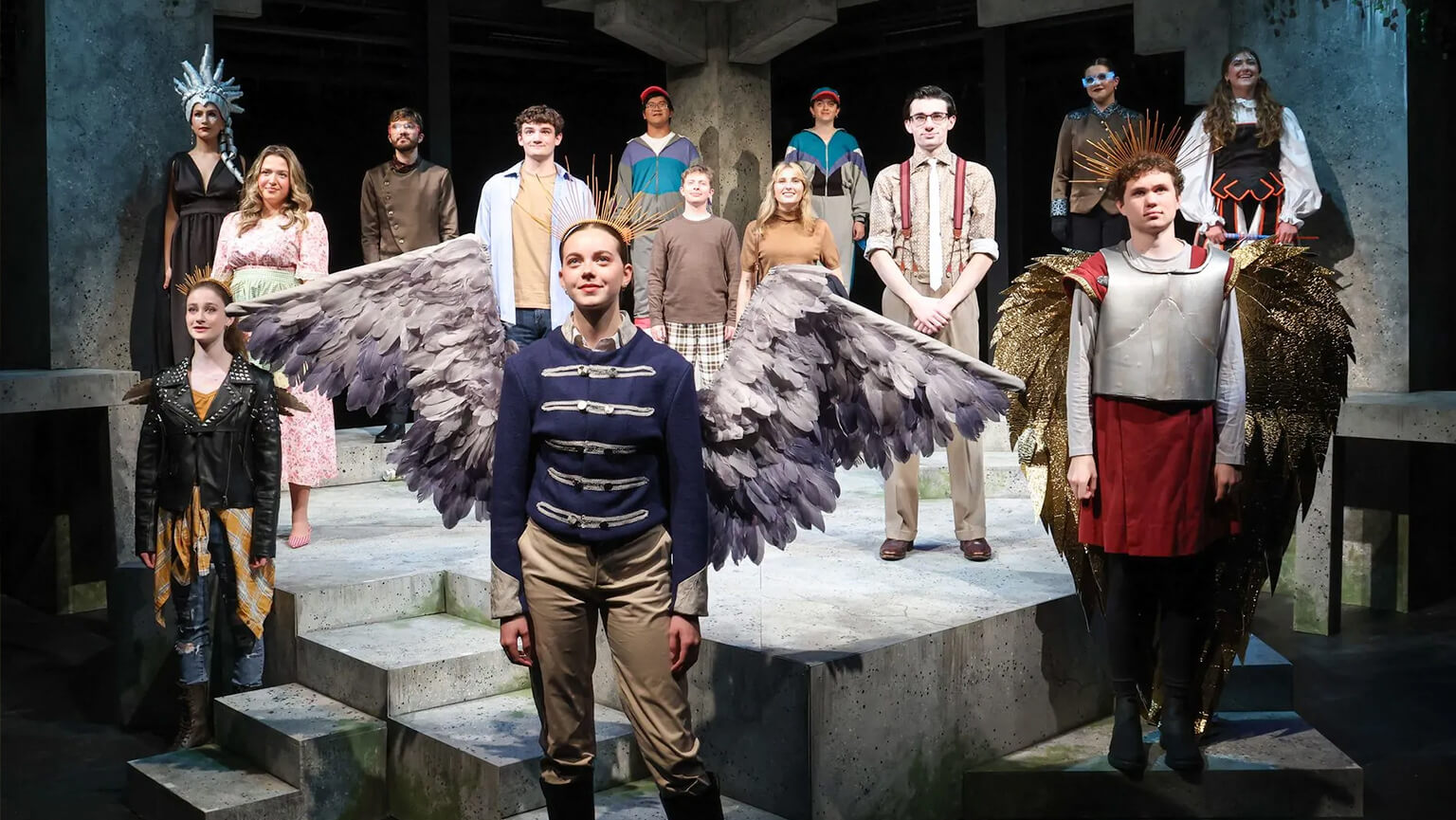 The cast of the play performs on stage, a person wearing colorful wings and showcasing their vibrant costumes.