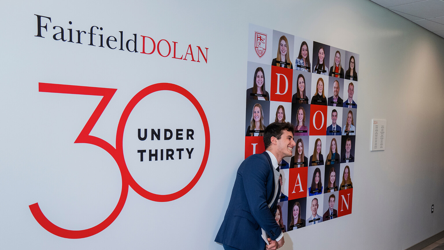 A student dressed in a suit stands before a wall with a prominent sign that reads "30 Under 30," celebrating young achievers.
