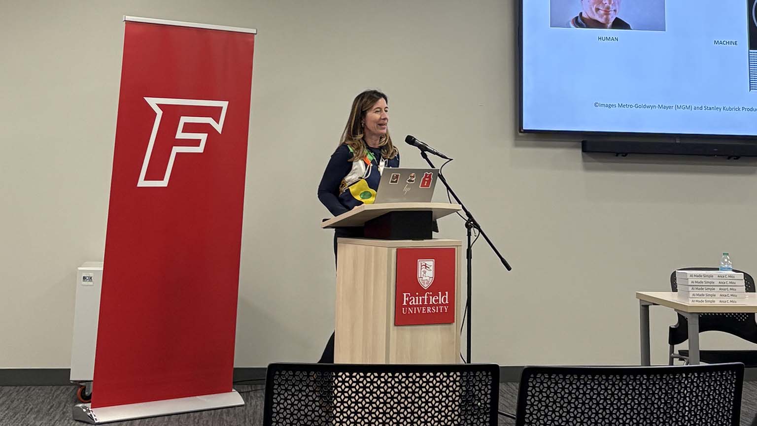 A woman stands at a podium, speaking in front of a large screen displaying information.