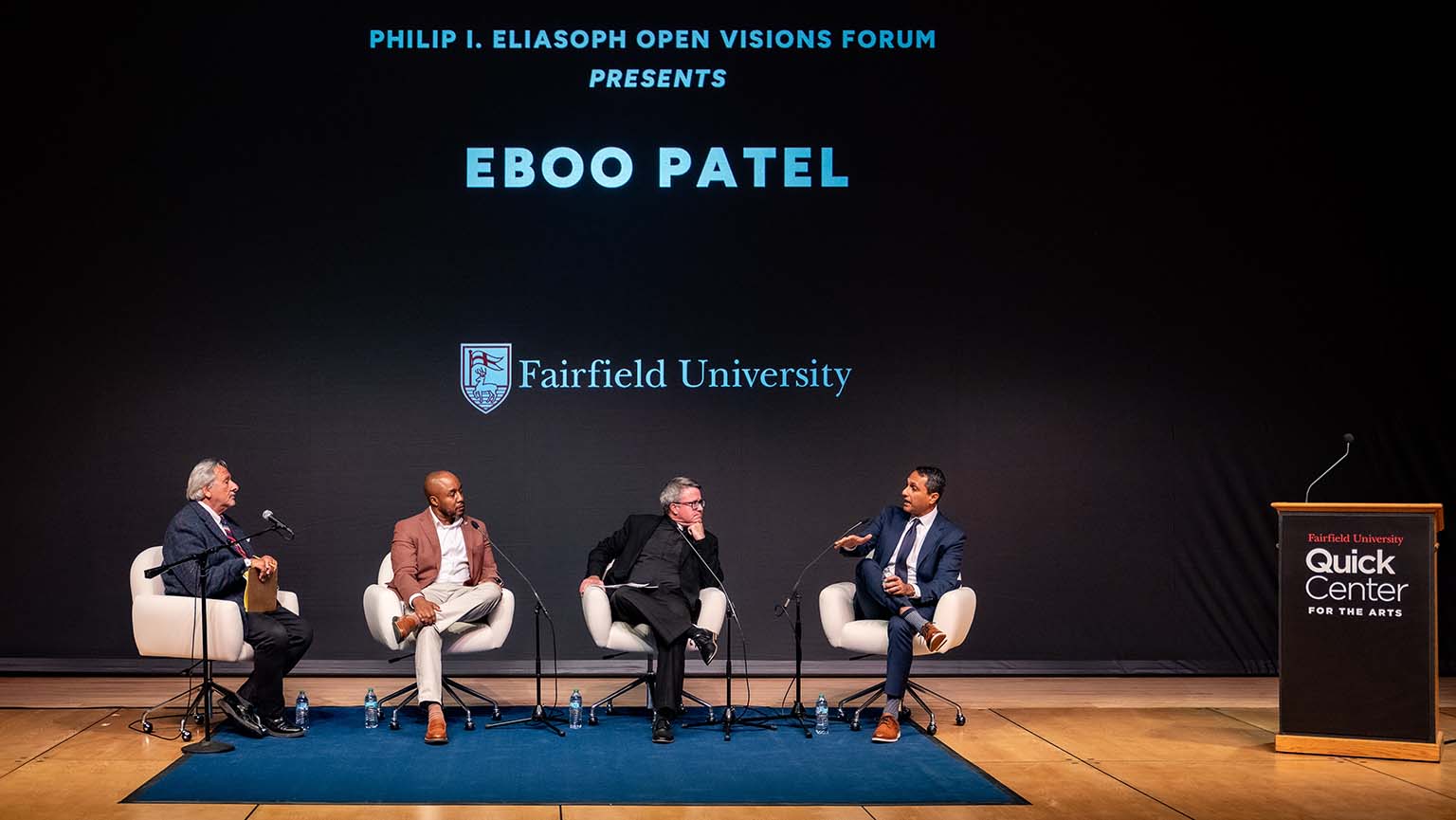Four men sitting on chairs, facing a stage, engaged in conversation.