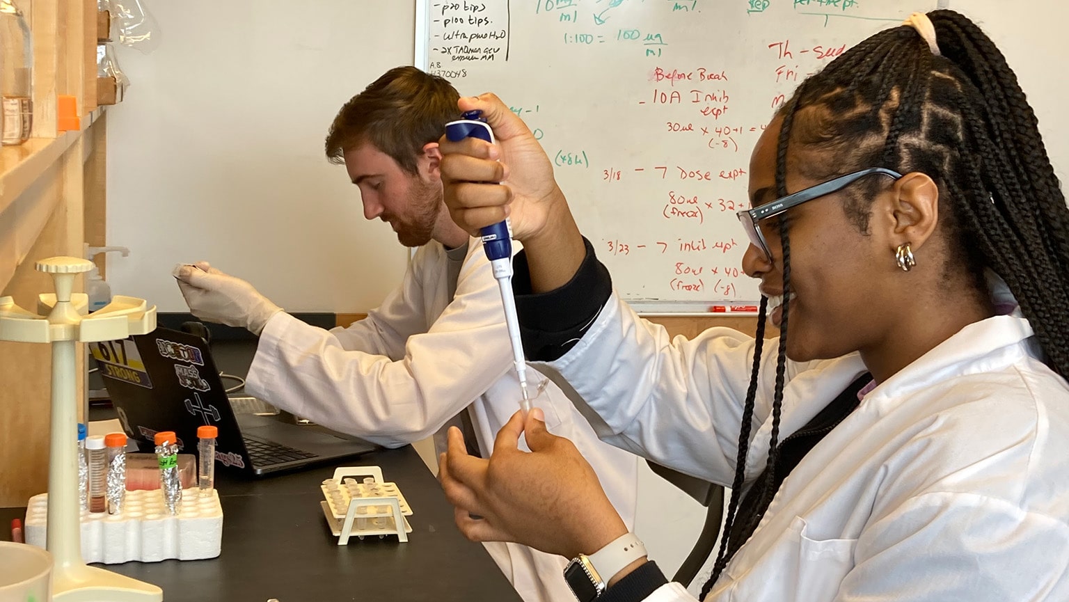 Two undergrad research students in lab coats pipetting samples into test tubes.