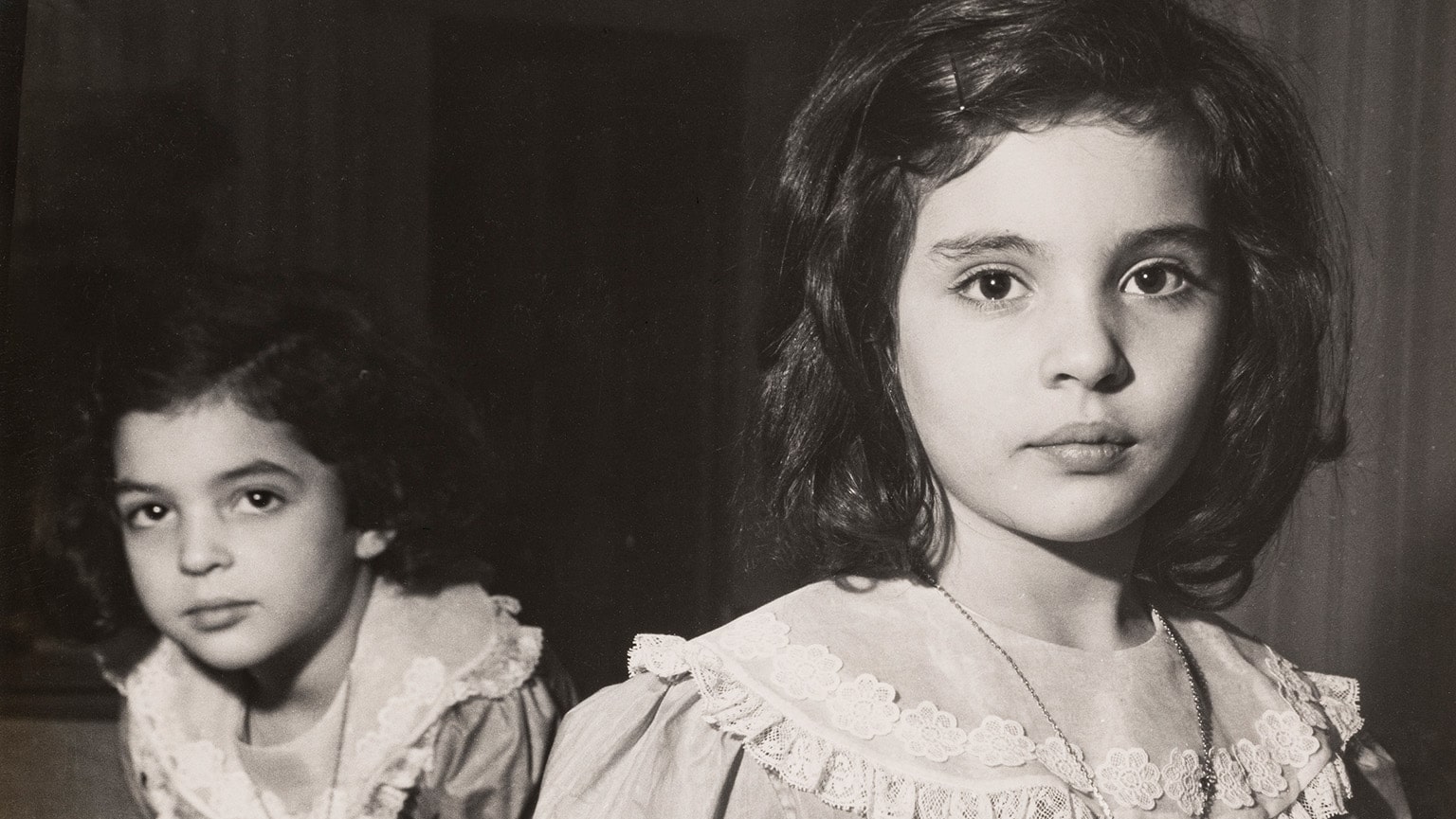 Historic black and white photo of two young girls in frilled dresses wearing heart necklaces.