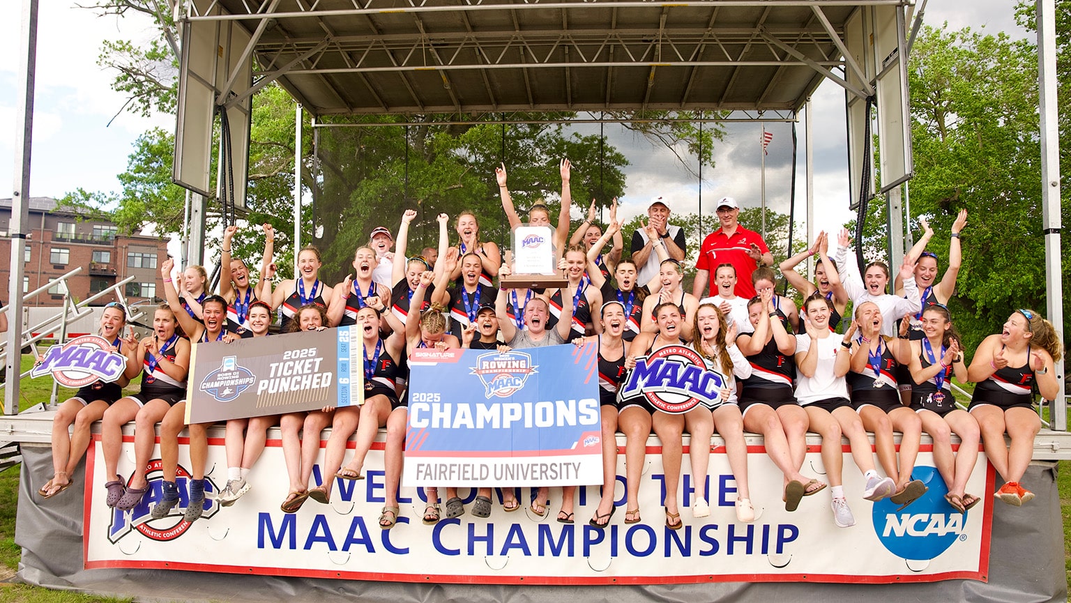 Fairfield's Women's Rowing team posing on a stage with their 2025 MAAC Championship winner banners, cutouts, and trophy.