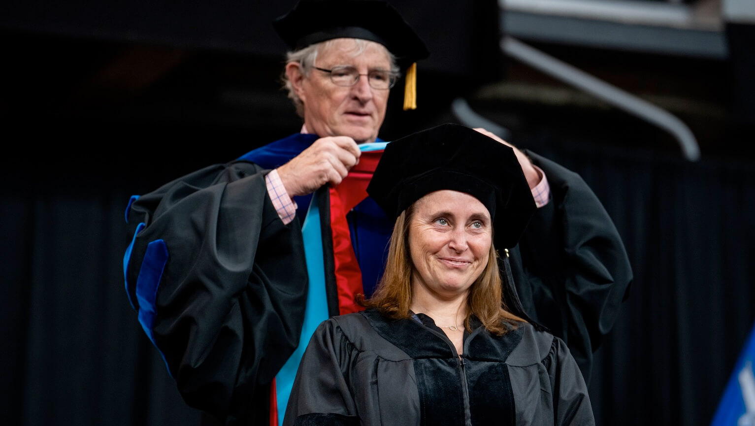 A woman in a graduation gown stands in front of a man wearing a graduation cap, both smiling proudly at their achievement.