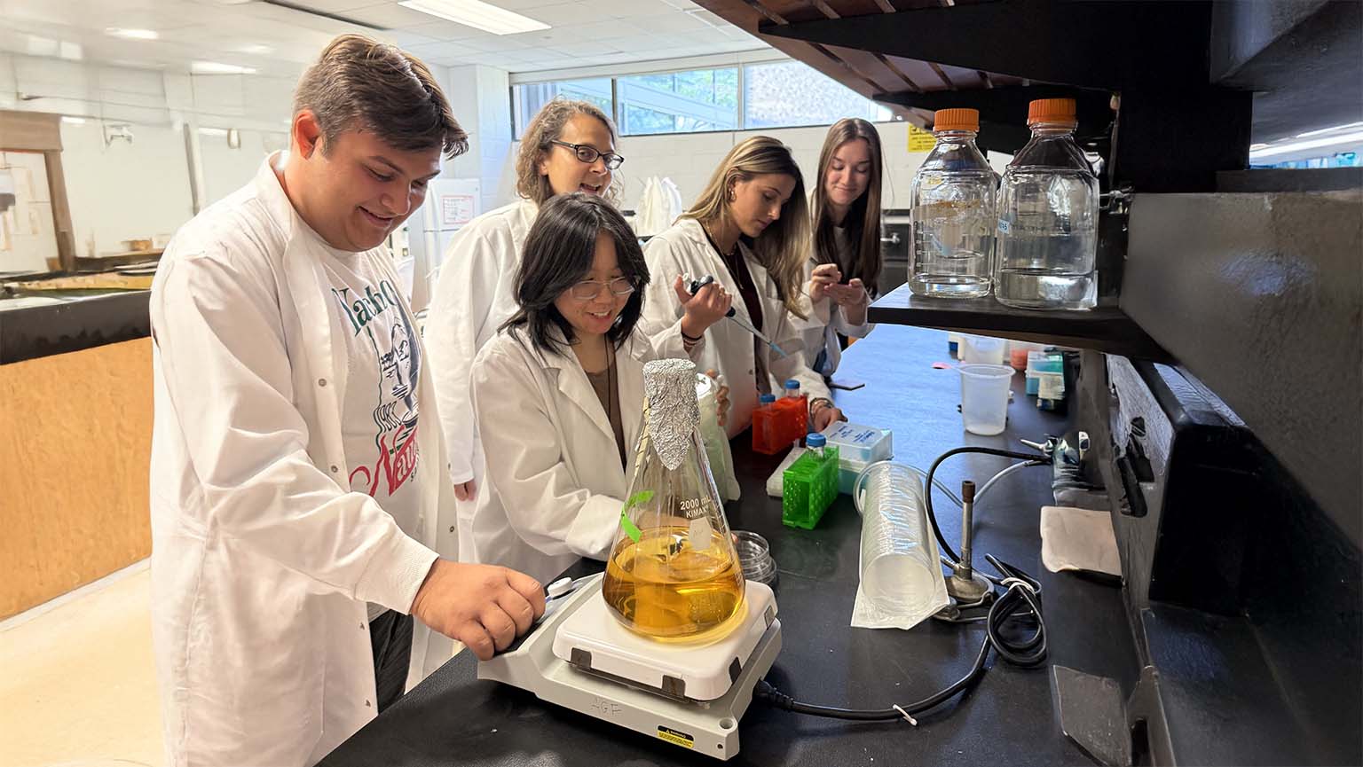 A group of students in lab coats collaborating on a science project at a lab table.