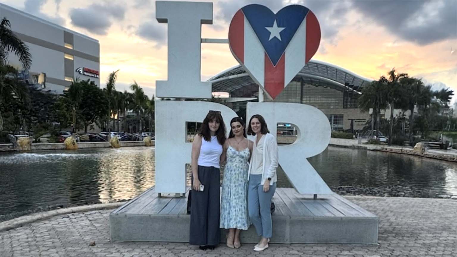 Three women stand in front of a large I ❤️ PR sign, with the heart displaying the Puerto Rican flag. A building, palm trees, and a pond are visible in the background at sunset.