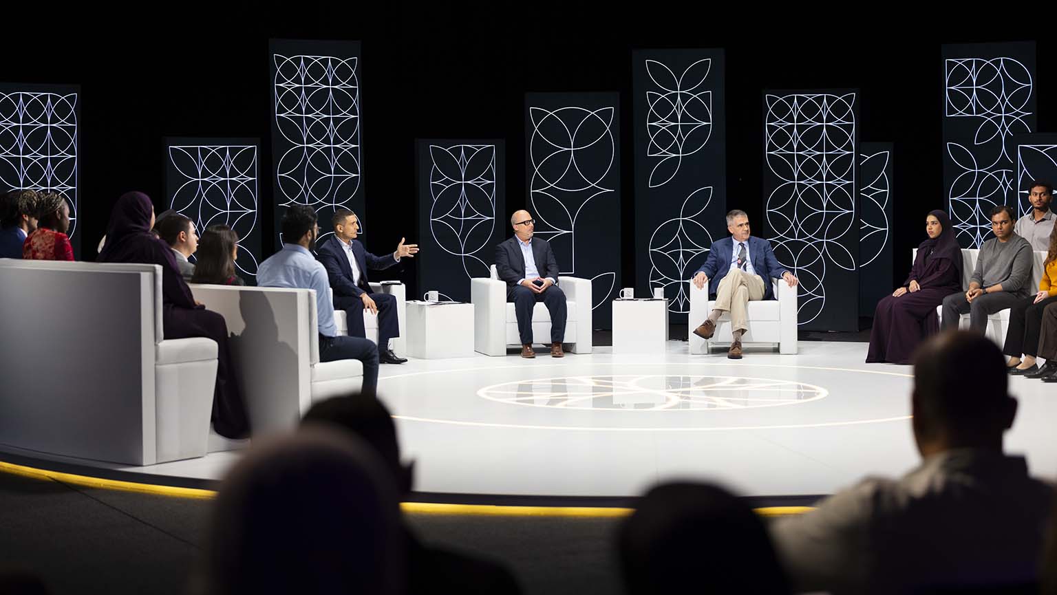 A group of people sit in a circular studio, engaged in a panel discussion. Three men sit on chairs in the center, while others sit on sofas around them. Decorative geometric panels are displayed in the background.
