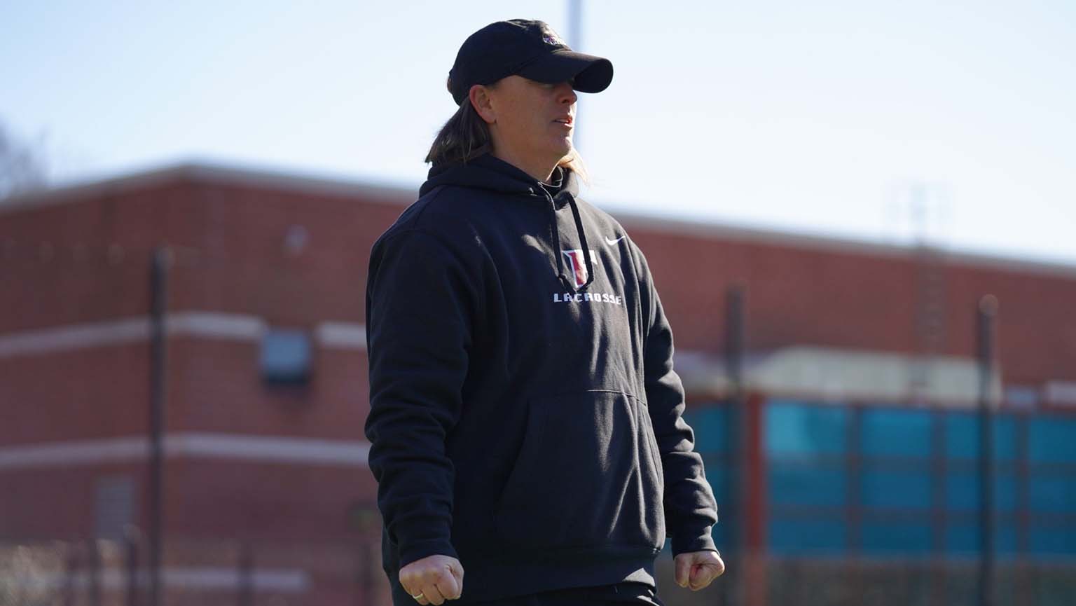 A person wearing a black cap and black hoodie stands outdoors in front of a blurred brick building, facing sideways with their fists clenched.
