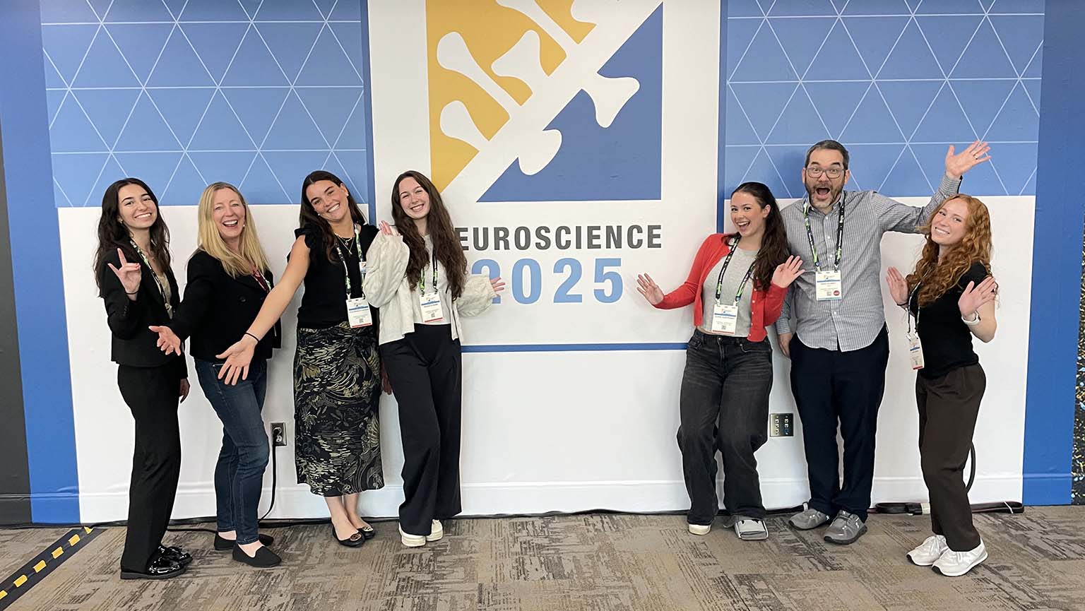 Seven smiling people stand in front of a wall with a “Neuroscience 2025” sign, posing with cheerful and welcoming gestures. 