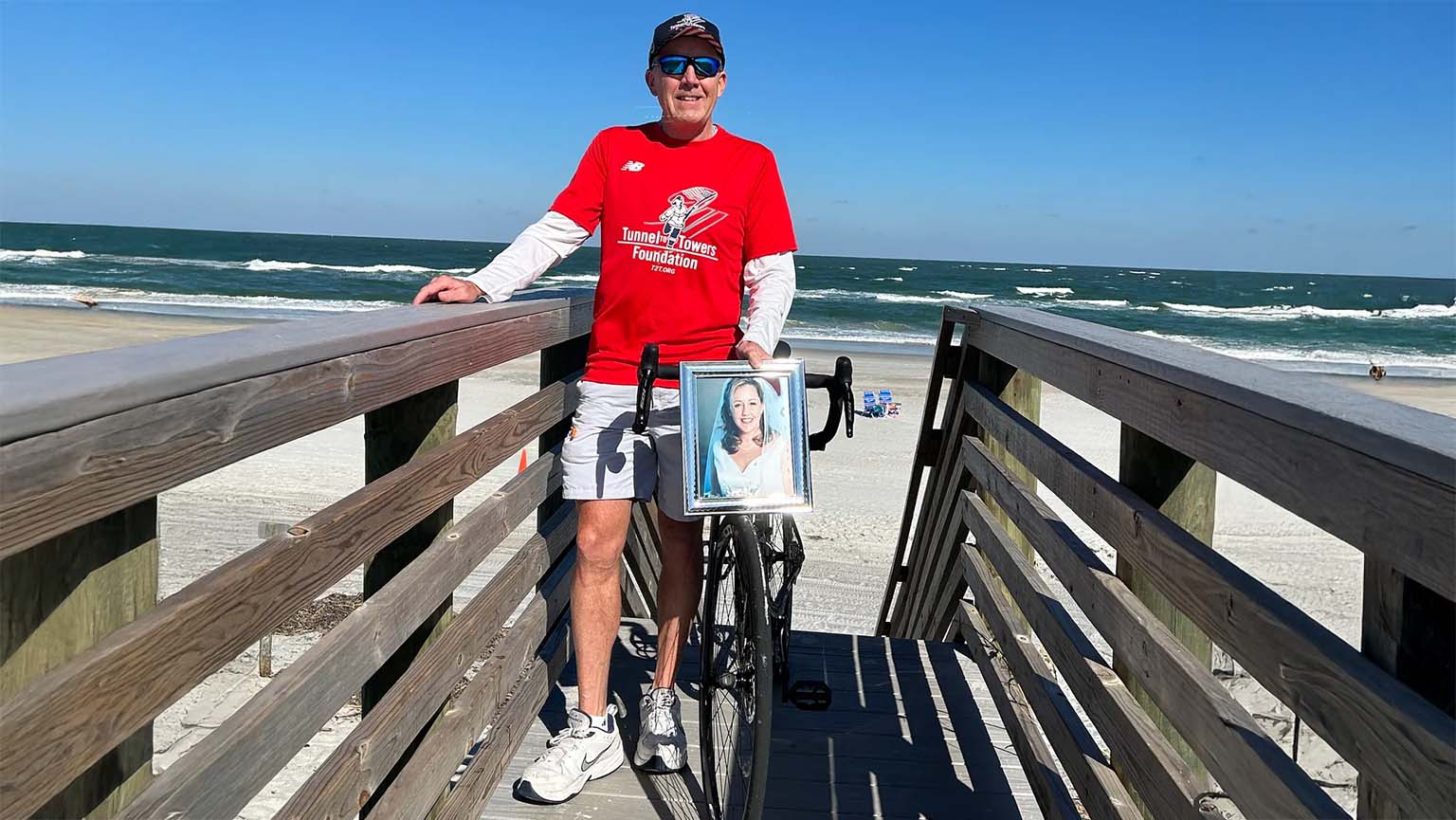 A man in a red shirt stands on a wooden boardwalk by the beach with a bicycle. Attached to the bike is a framed photo of a woman. Ocean waves and a clear blue sky are visible in the background.