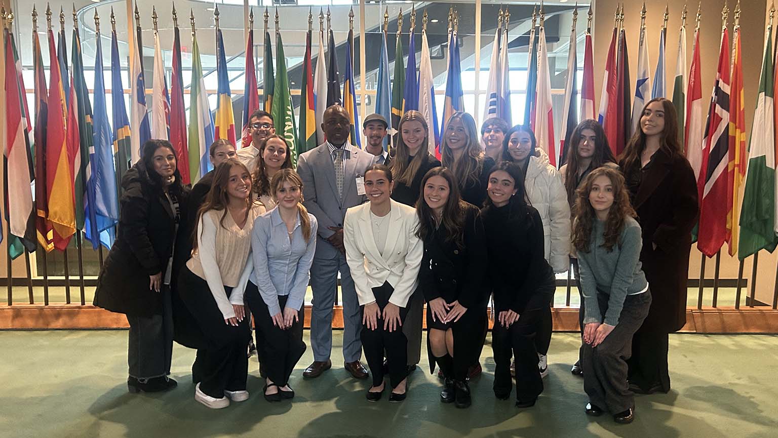 A diverse group of young adults and one man in a suit pose together in front of a display of various international flags inside a formal building.