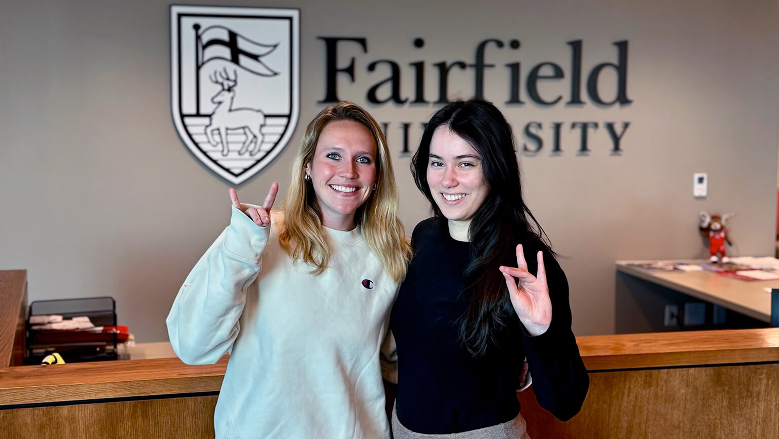 Two female friends smile and gesture in front of a "Fairfield University" sign. The scene is warm and welcoming, with a university emblem in the background.