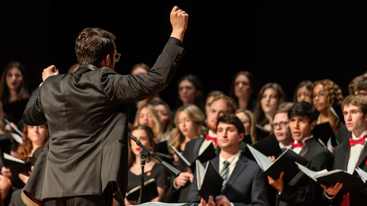 Director of the Fairfield University Glee Club, professor Michael A. Ciavaglia wears a black suit and conducts a choir of diverse singers holding music sheets on stage. The singers, wearing formal attire, focus intently.
