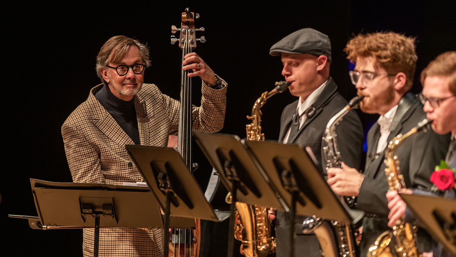A jazz band performs on stage with bassist Brian Q. Torff, Fairfield University professor of music, in a checkered jacket, alongside saxophonists in suits. The mood is lively and focused, with music stands in view.