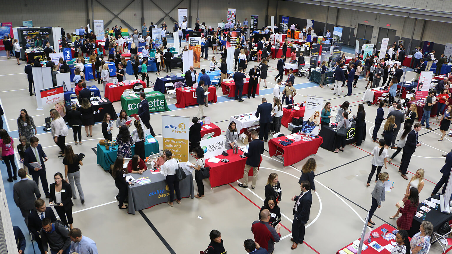 A bustling job fair in a large gym with rows of tables covered in colorful cloths. People engage in discussions, creating a lively, professional atmosphere.