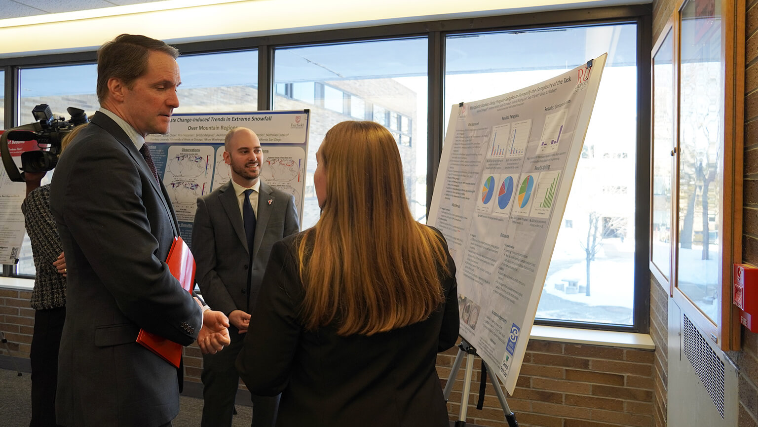 U.S. Representative Jim Himes and students in business attire stand near a large poster board displaying charts and graphs. A snowy scene is visible through the windows behind them.