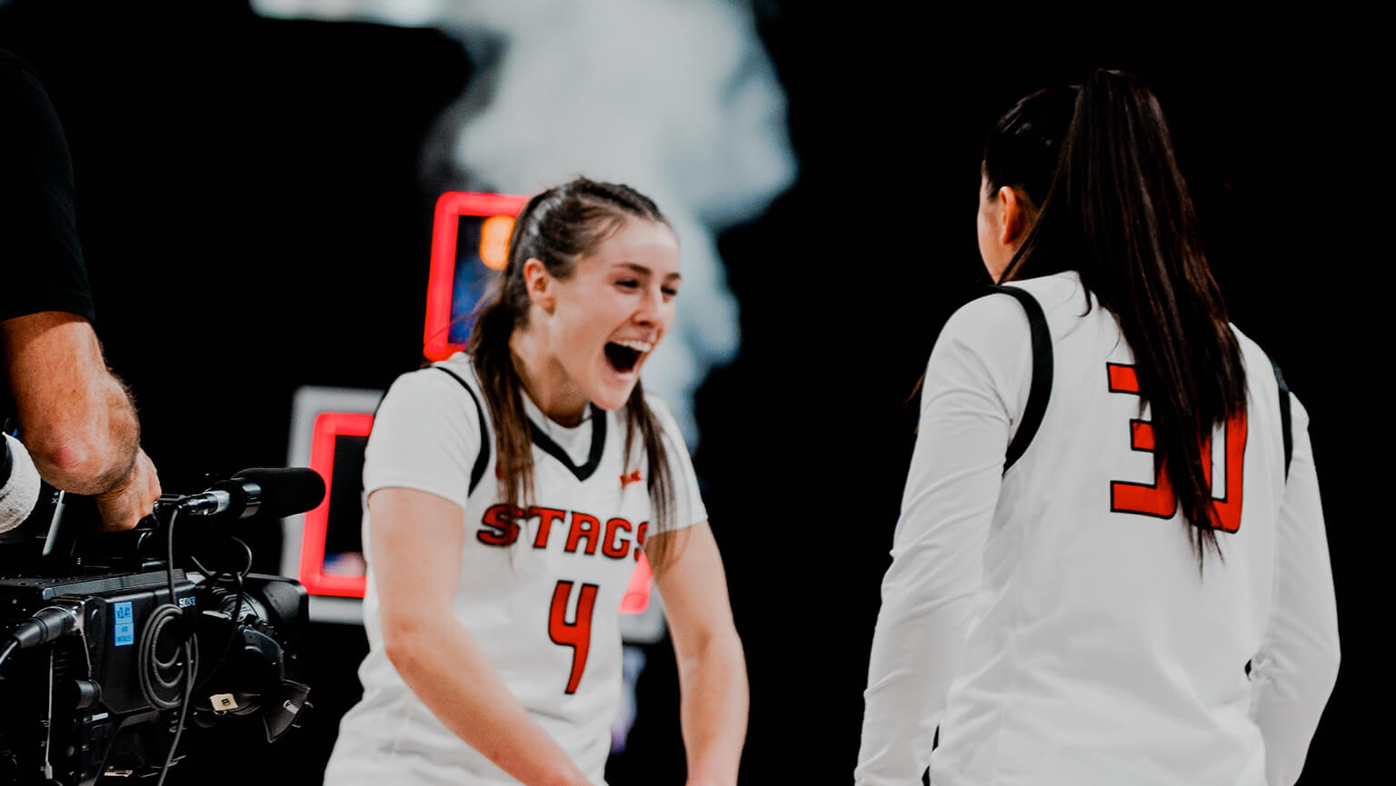 Kaety L’Amoreaux ‘27 and basketball teammate in white jerseys with red numbers celebrate energetically on a court. A camera operator films them, and digital screens are blurred in the background.