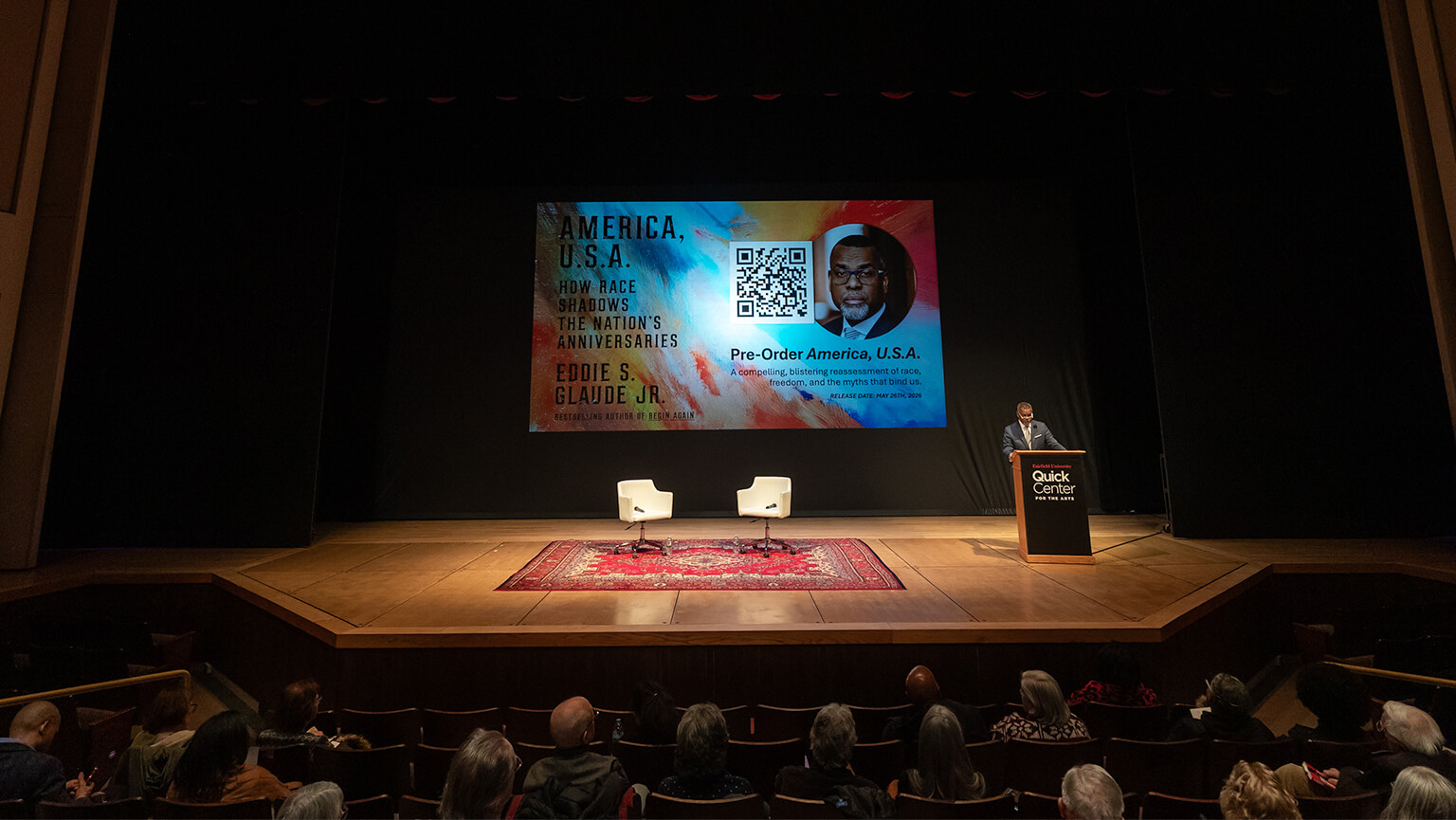 Eddie S. Glaude Jr., PhD speaking at a podium on a stage with two empty white chairs and a red rug. A large screen displays a colorful book cover and a QR code, watched by an audience in a dimly lit auditorium.