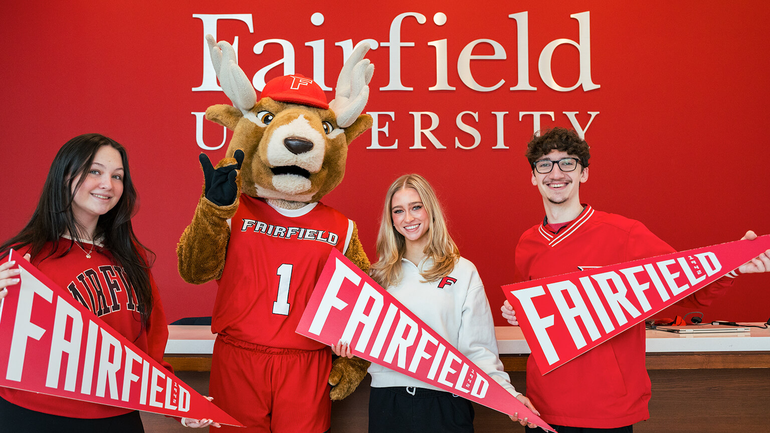 Three students and the Lucas the Stag mascot hold Fairfield University pennants, standing in front of a red wall with the university's name, smiling and cheerful.