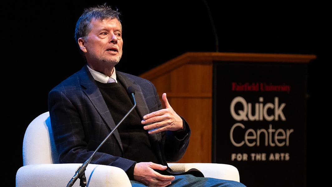 American filmmaker Ken Burns giving a lecture at Fairfield University while seated casually on stage.