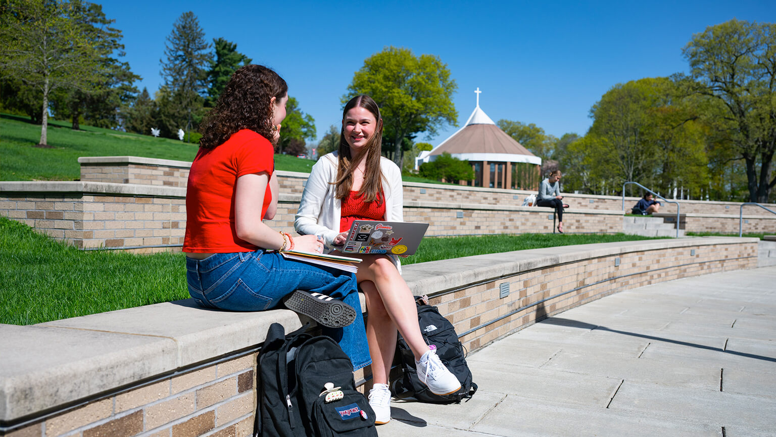 Two female students sit on a brick wall in a sunny part of Fairfield University campus, talking and smiling, with laptops and backpacks. A small chapel is visible in the background.