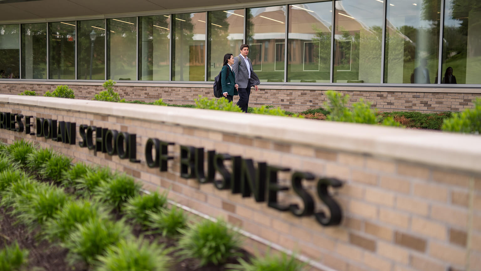 Two students walking in front of the Charles F. Dolan School of Business. The building features large glass windows and well-kept landscaping.