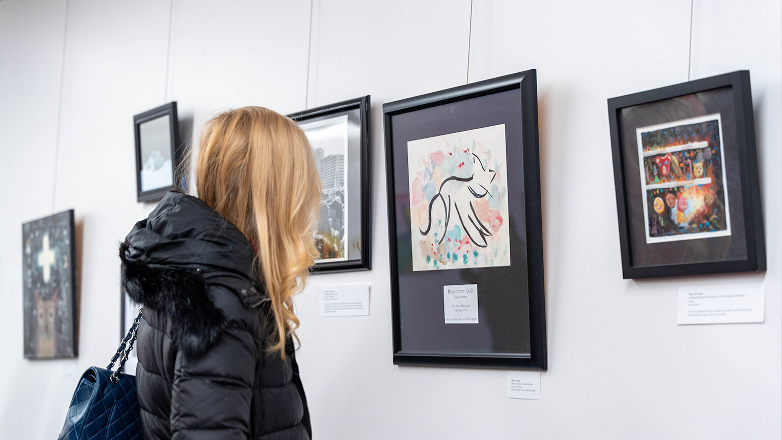 A woman with long blonde hair in a black coat admires framed abstract artwork in a gallery. The scene conveys a sense of curiosity and appreciation.