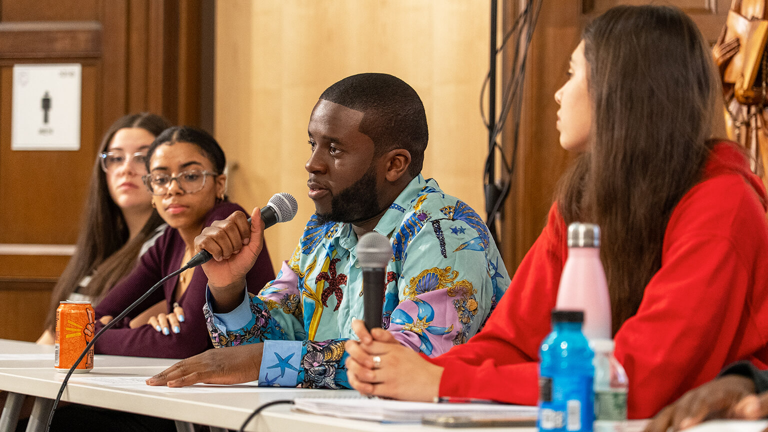 A diverse panel of people at a discussion table. A man in a colorful shirt speaks into a microphone, engaging the attentive audience.