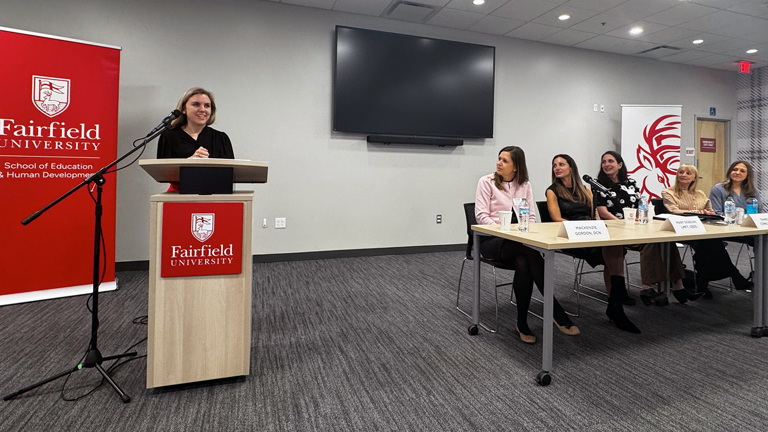 A woman speaks at a podium with a Fairfield University banner. Women sit at a table, smiling, in a conference room setting. The tone is professional and engaging.