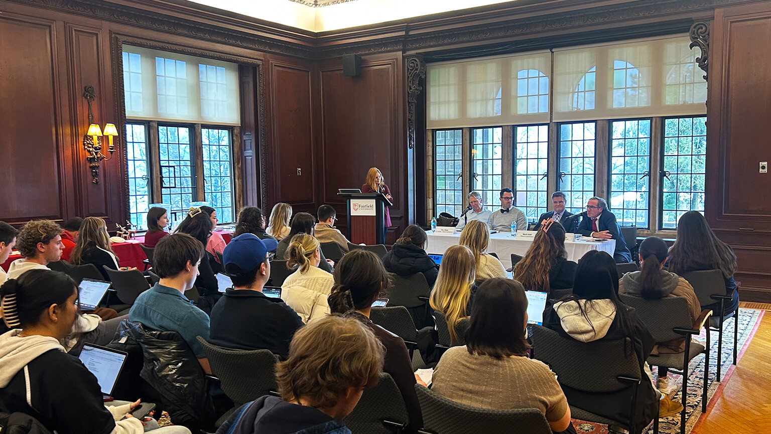 A panel discussion in a wood-paneled room, with people seated facing four speakers at a table. A woman stands at a podium, and the mood is attentive.