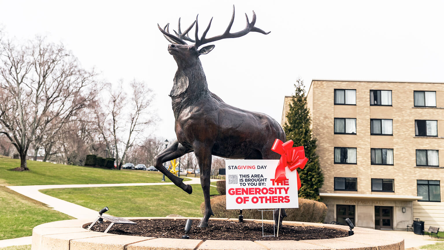A statue of a rearing stag with antlers on Fairfield University campus, a sign at its base reads, "This area is brought to you by the generosity of others," with a red ribbon.