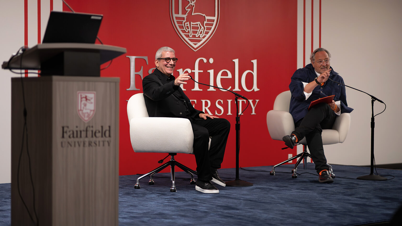 Stephen Wilkes and Philip I. Eliasoph sit on stage at Fairfield University, smiling and engaging with the audience. One holds a microphone. The atmosphere is lively and conversational.