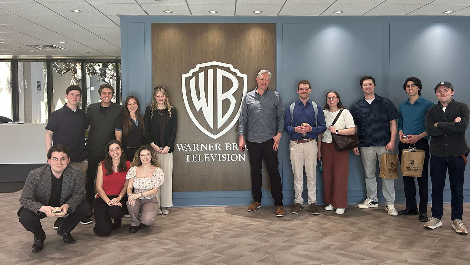 A group of Fairfield students, smiling and casually dressed, stand in a modern lobby with a large Warner Bros. Television logo on the wall behind them.
