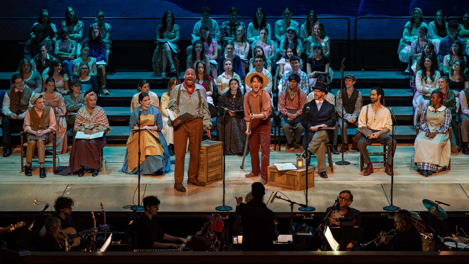 A diverse group of people performing on stage with an orchestra in the background, creating a lively musical atmosphere.