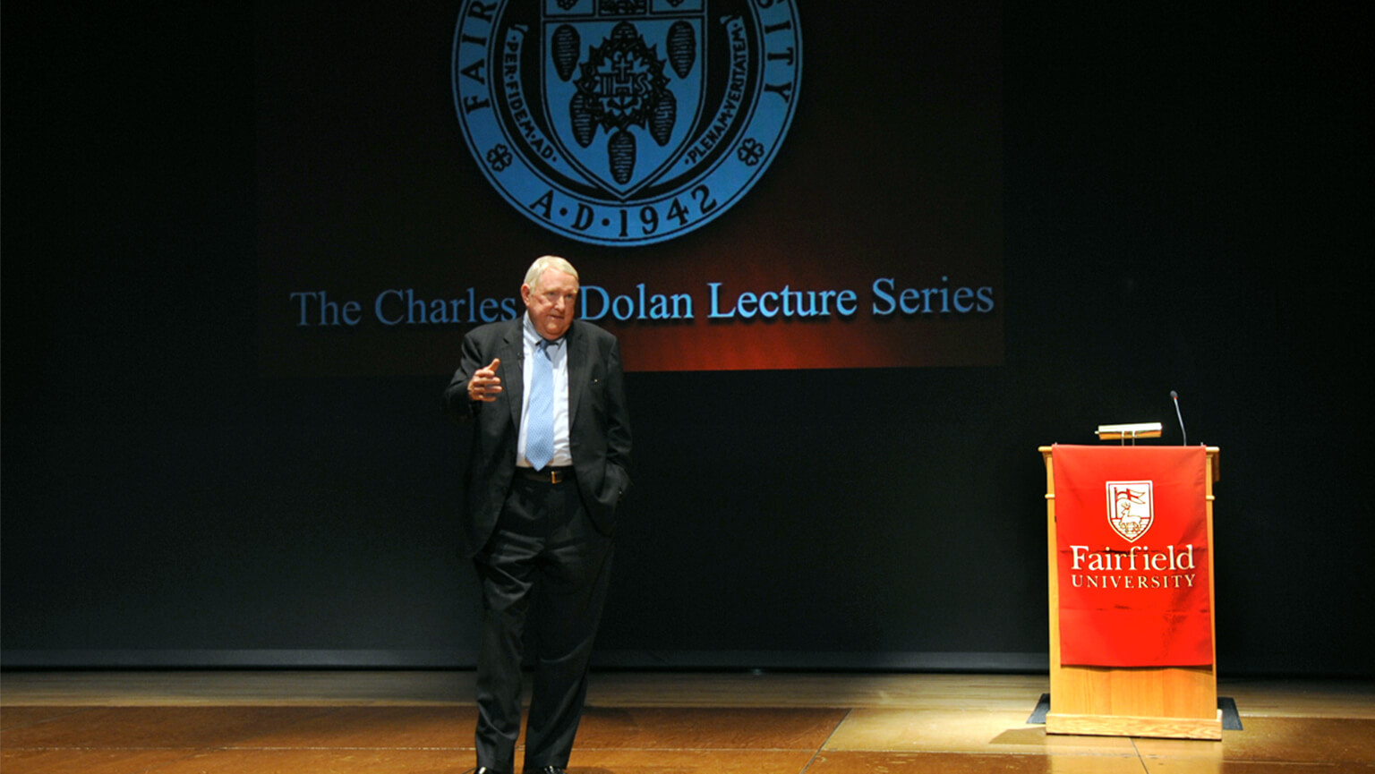 E. Gerald Corrigan ’63, PhD in a suit gestures passionately while speaking at a podium. Behind him, partially visible text reads "Lecture Series," indicating a formal setting.