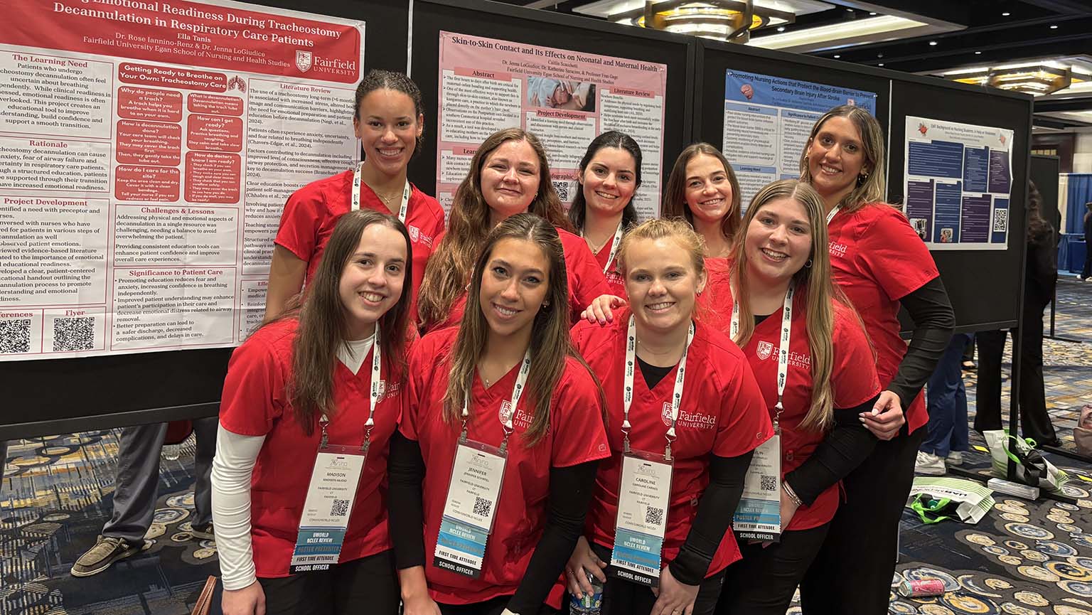A group of young women in red shirts stands beside a colorful poster, smiling and engaging with each other.