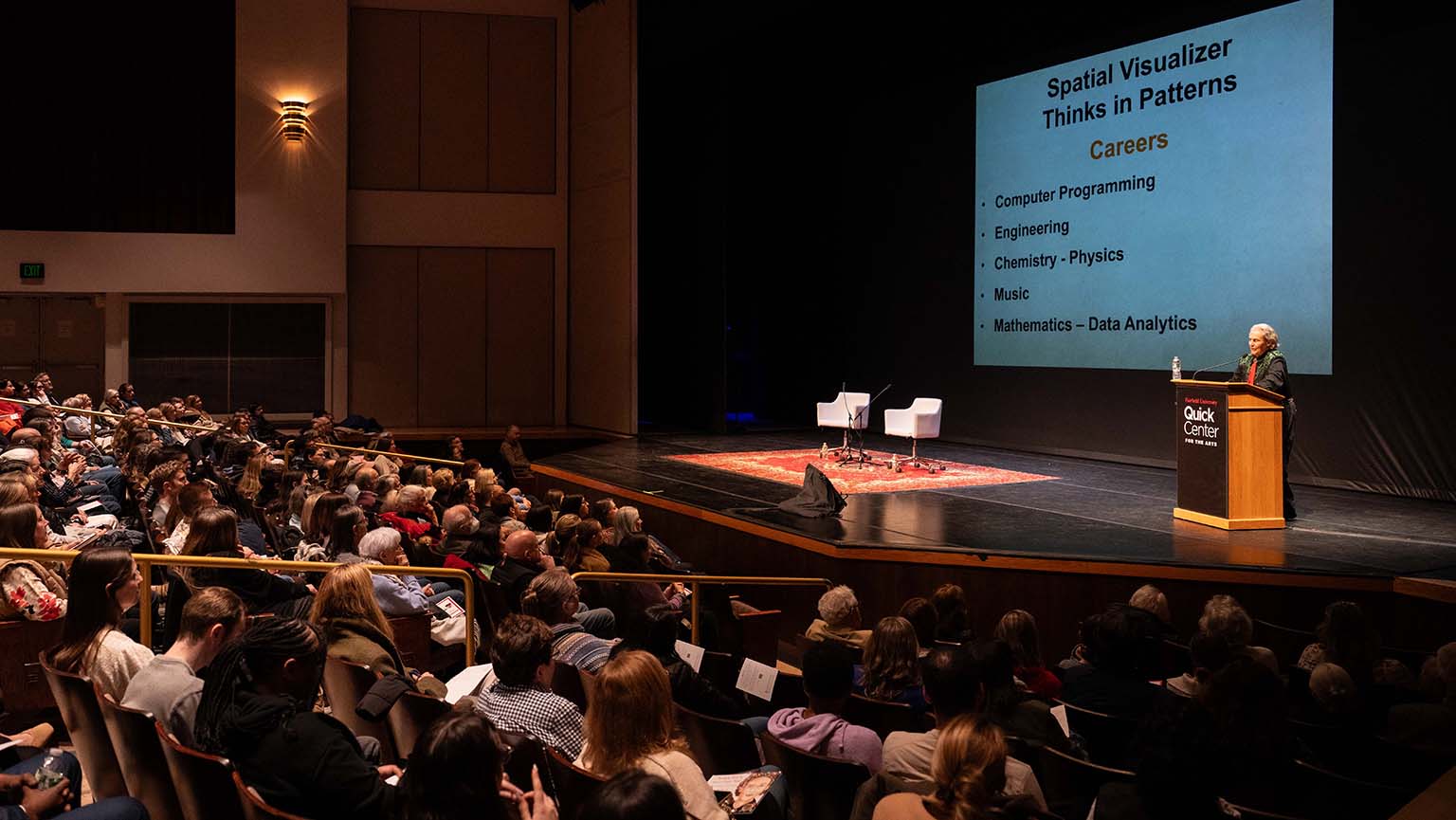A speaker addresses a large crowd in an auditorium, engaging the audience with his presentation.
