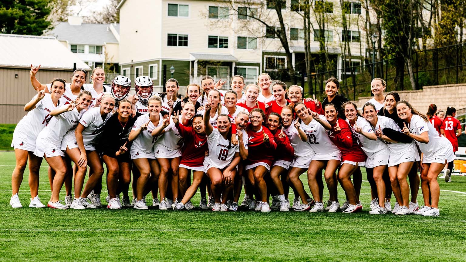 The women's lacrosse team stands on the field, united and smiling for a team photo.