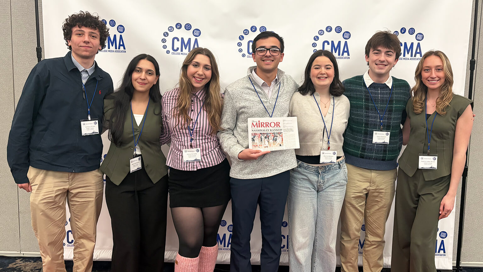 Students stand smiling in front of a CMA backdrop. One holds a Newspaper reading "MIRROR." They appear happy and proud, suggesting a celebratory event.