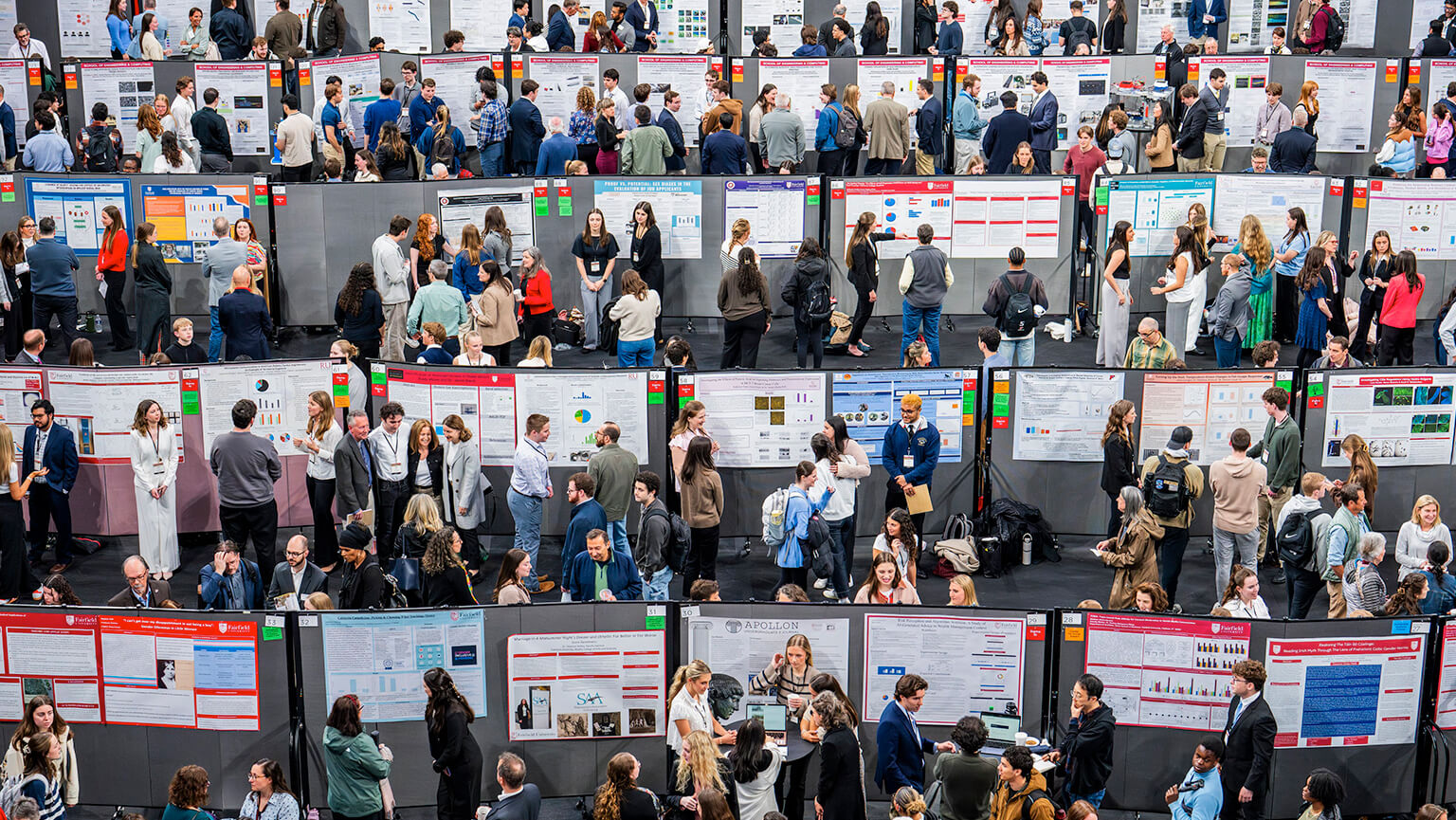 A crowded research symposium session with diverse groups engaging in discussions around various research posters. The atmosphere is active and collaborative.