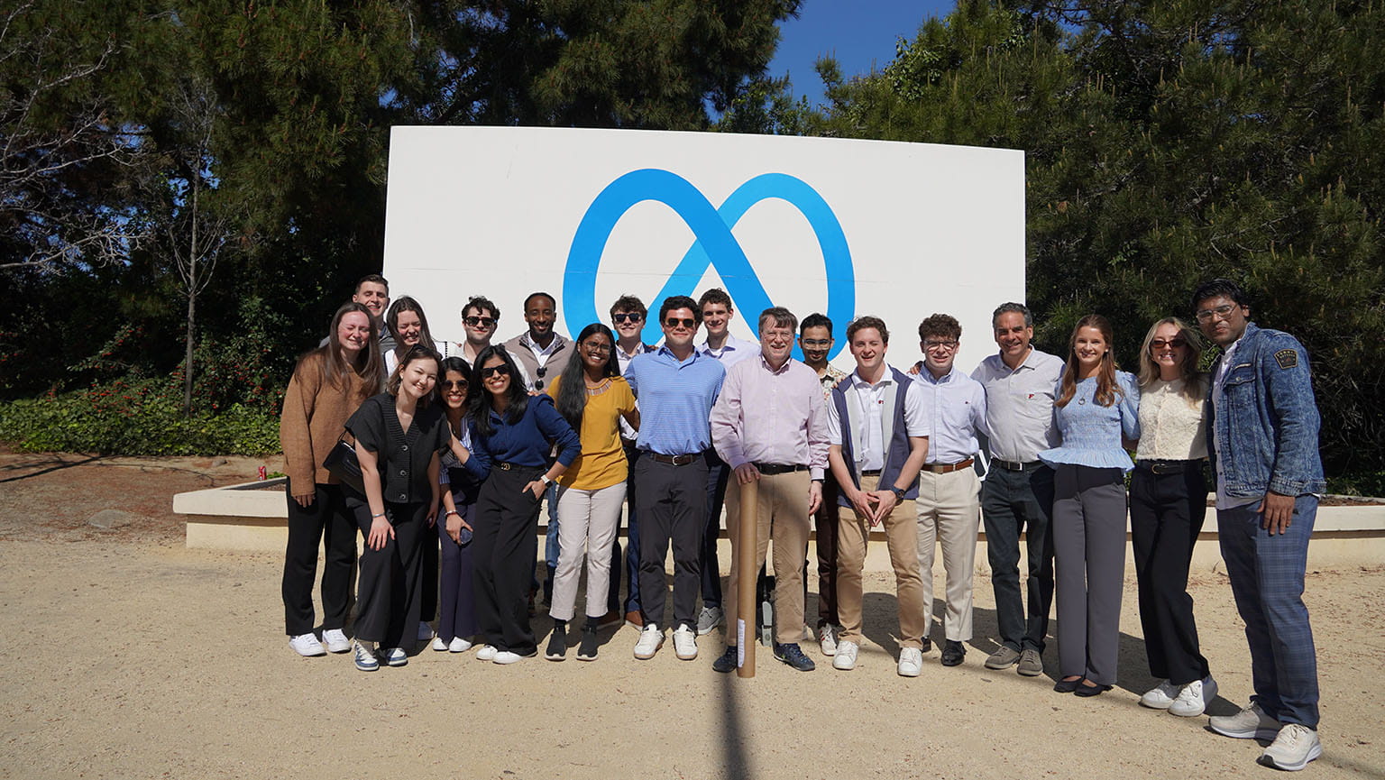 A group of people stand smiling in front of a large Meta logo sign outdoors, surrounded by trees. They appear to be casually dressed in a friendly setting.