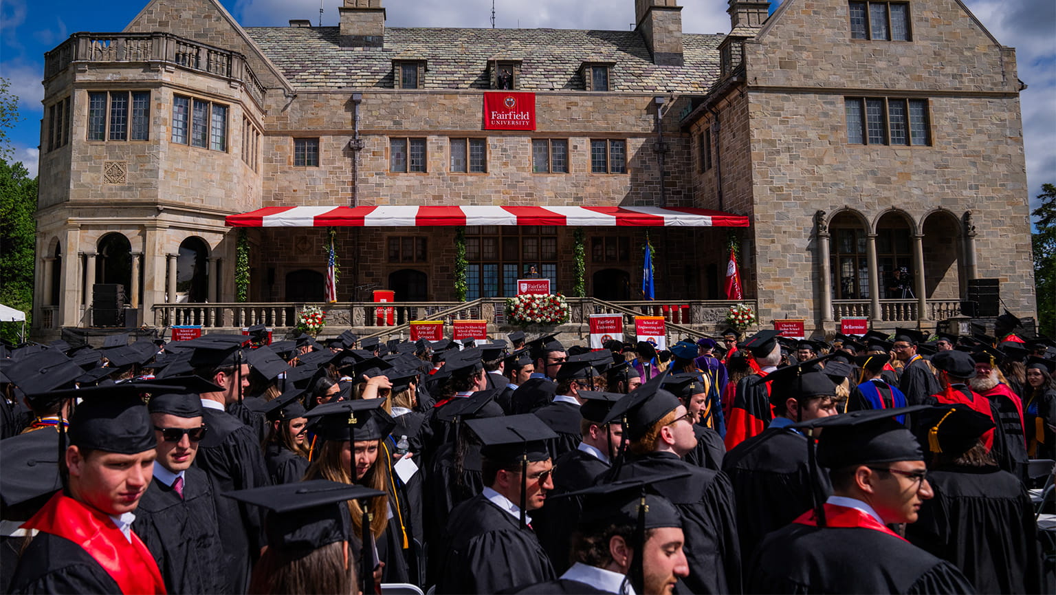 Students in caps and gowns lined up at their seats on a bright, sunny day as Commencement exercises begin.
