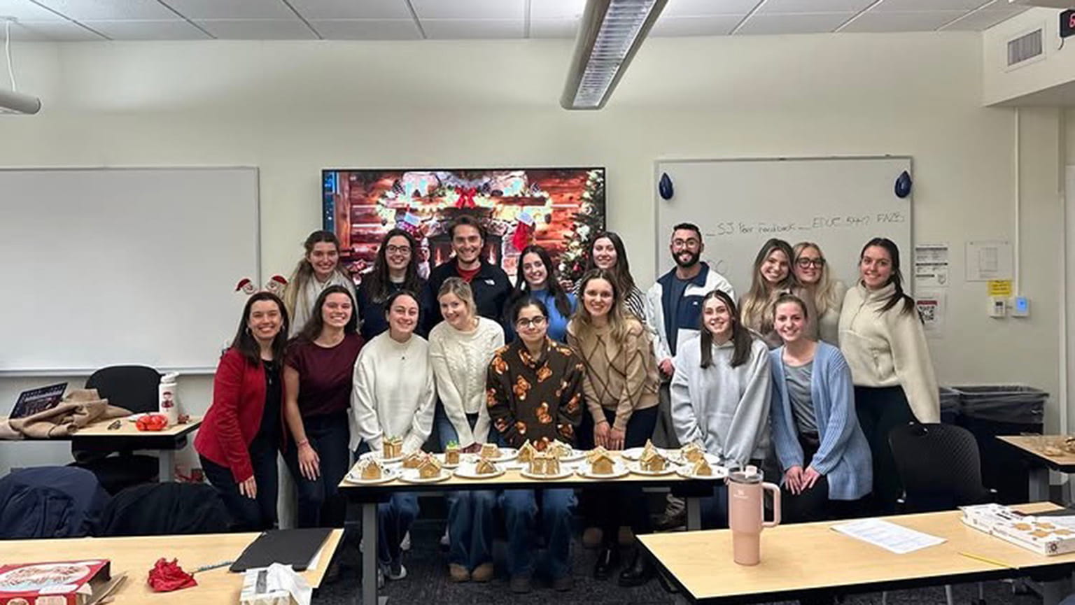 A group of smiling people stand in a classroom, gathered around a table with gingerbread houses. The room has a festive, holiday feel.