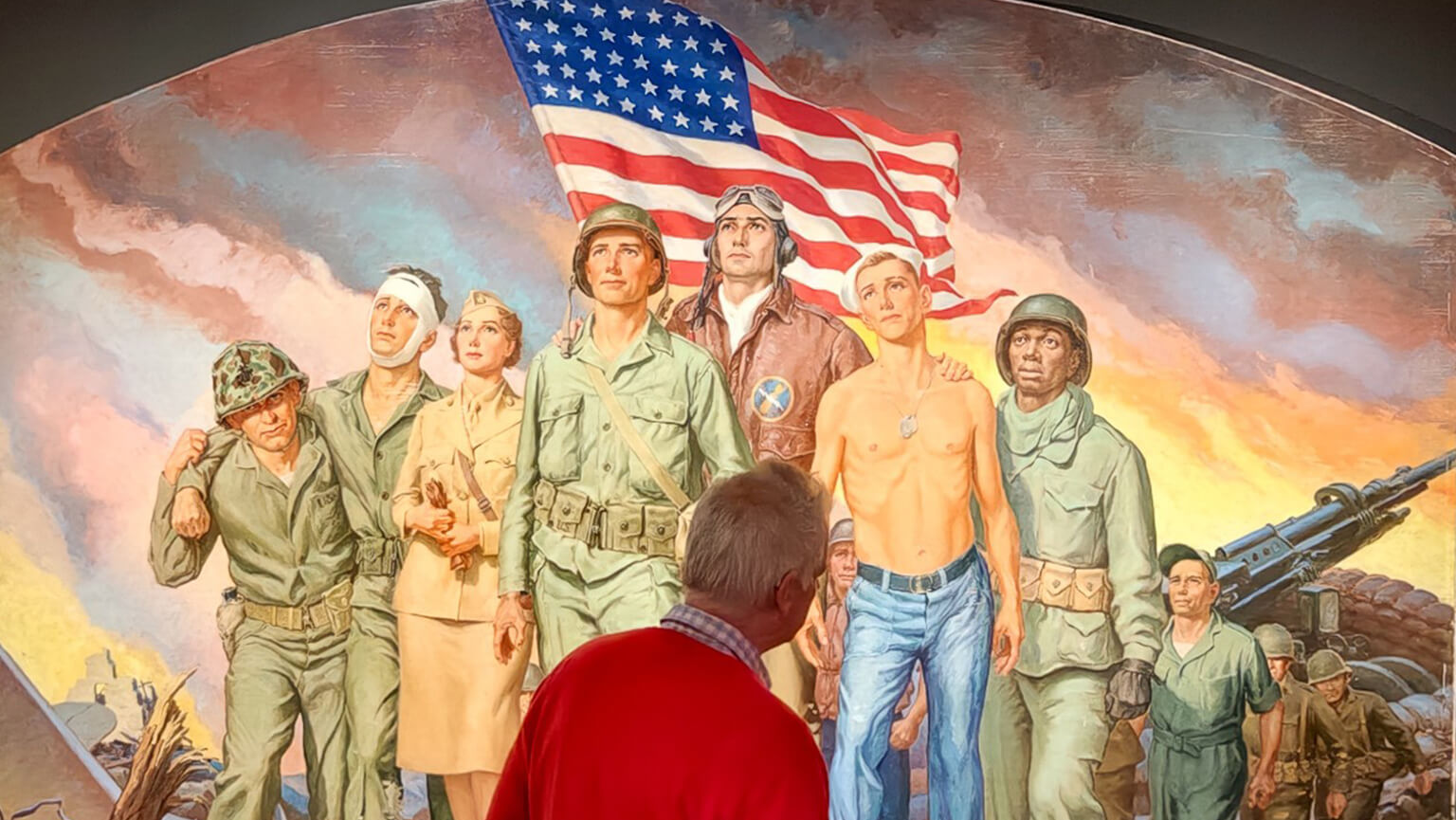 A person in a red sweater stands in front of a colorful mural, Robert Lynn Lambdin’s 1958 [Heroes of World War II] painting, in Meditz Gallery depicting diverse soldiers and a nurse, with an American flag in the background.