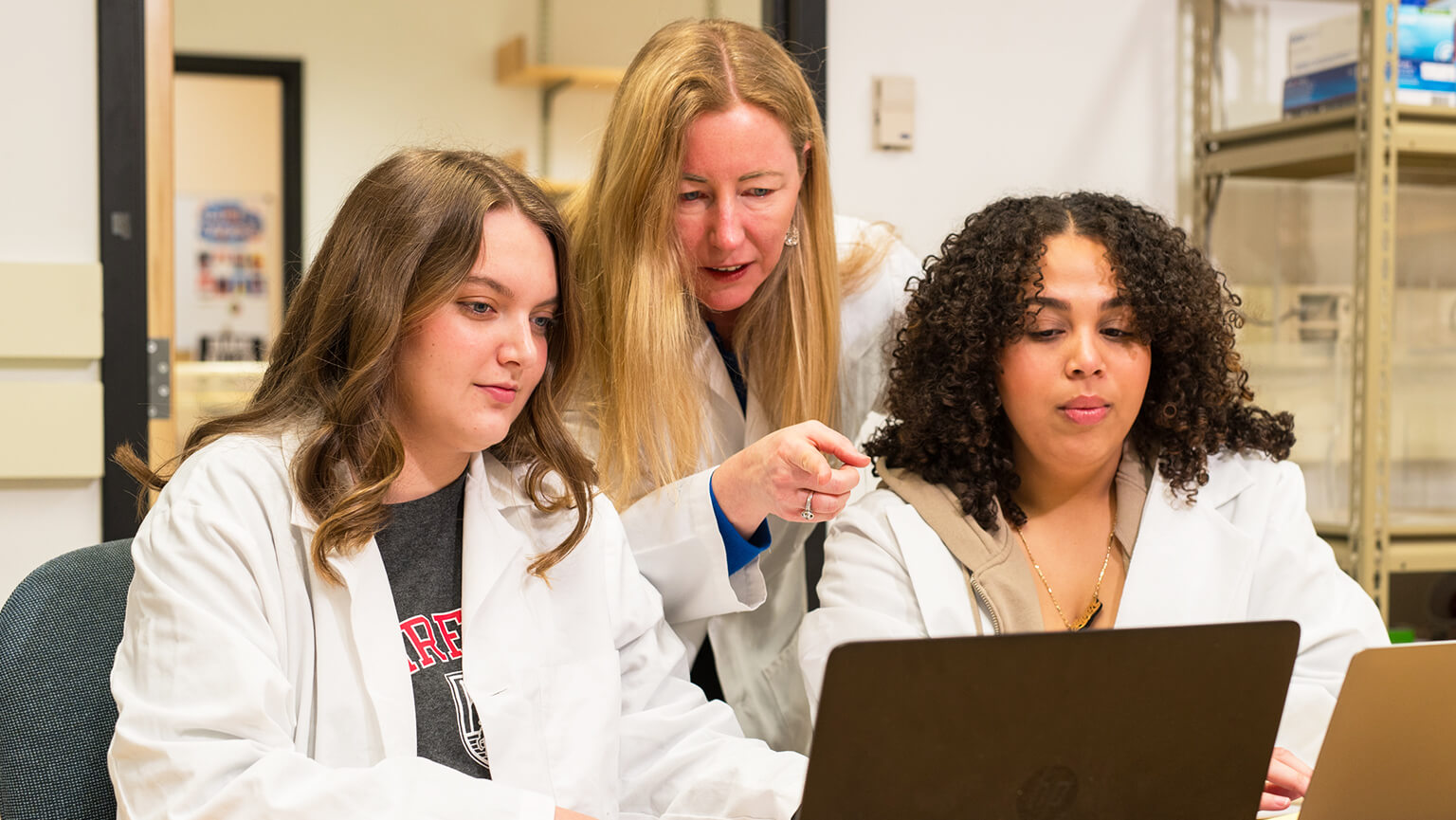 Three women in lab coats collaborate in a lab, focusing on laptops. The atmosphere feels studious and engaged, emphasizing teamwork and learning.