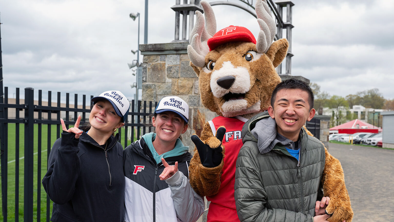 Three smiling students pose with Lucas the Stag mascot. They stand outdoors near a fence, exuding a cheerful and playful vibe.