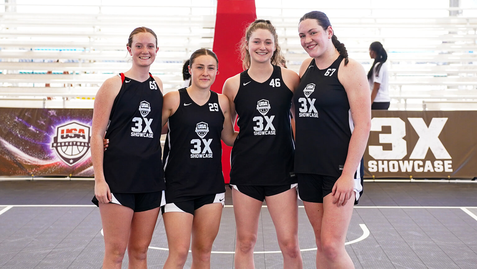 Four smiling young women in matching black basketball jerseys stand on an indoor court with "3X Showcase" branding in the background.