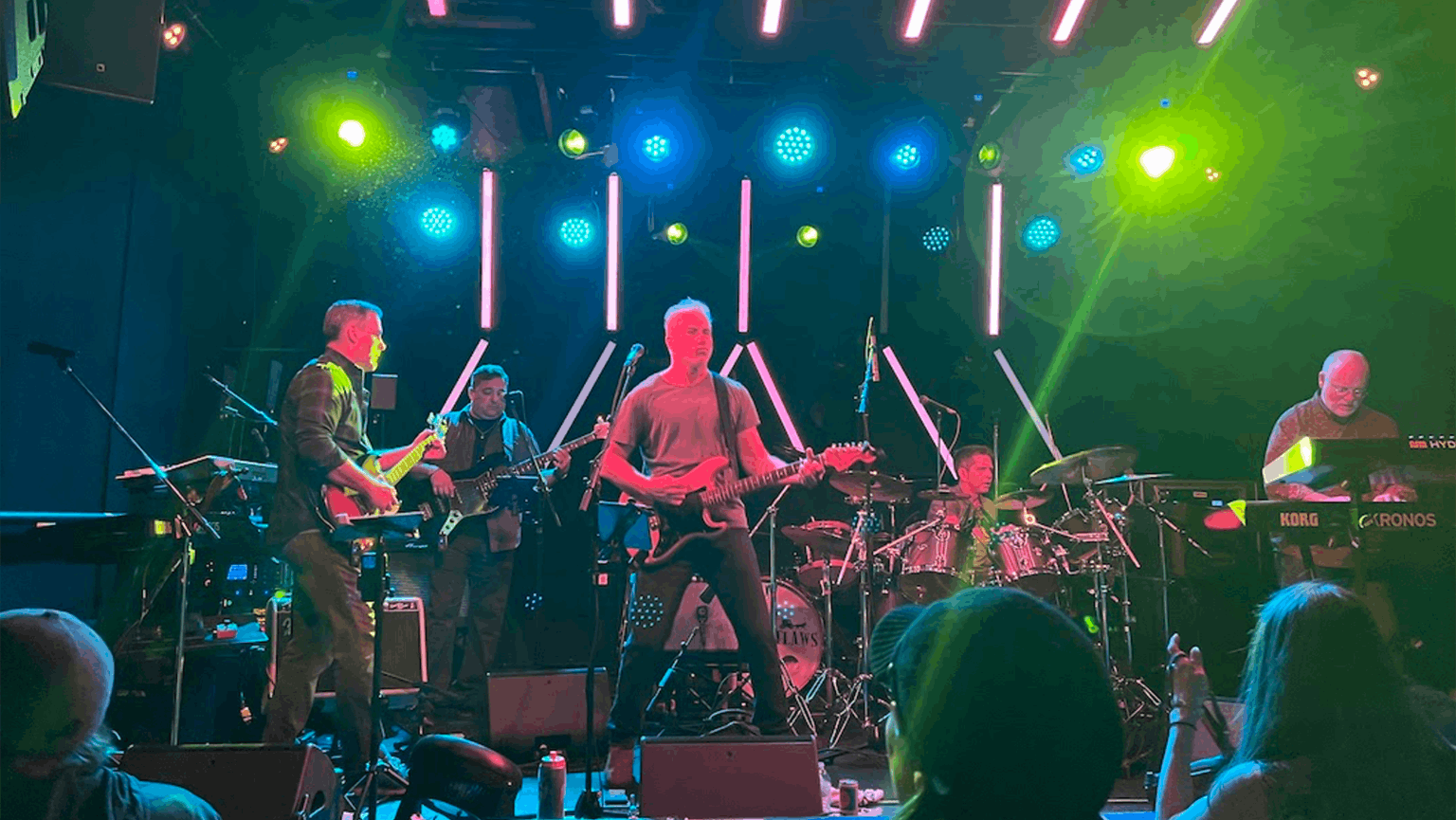 A band performs on stage under colorful lights, with musicians playing guitar, bass, drums, and keyboard while an audience watches.