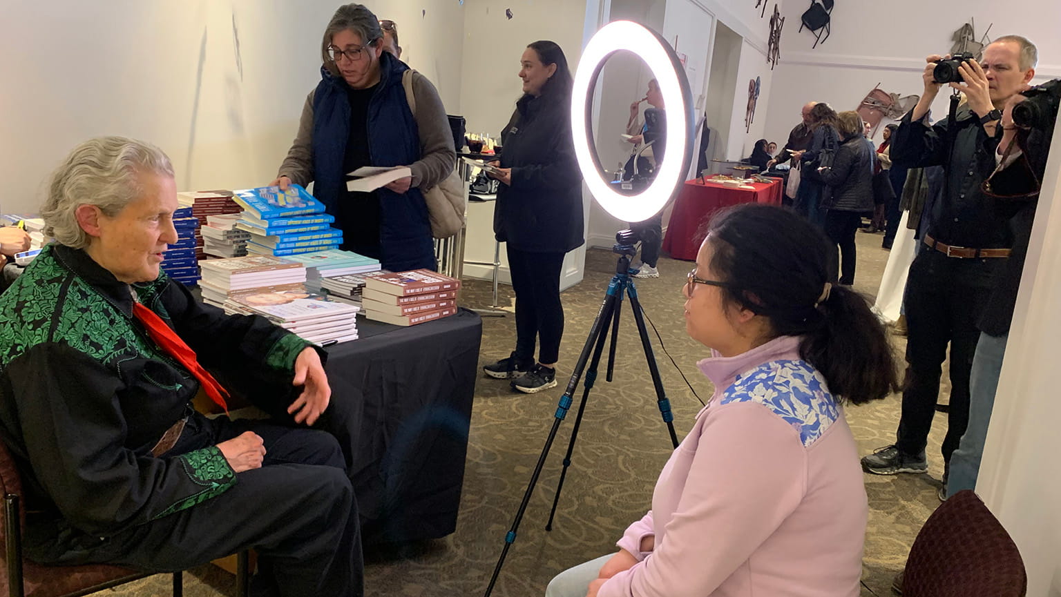 Two people are seated and talking in front of a ring light and stacks of books while others stand and a photographer takes pictures in an indoor event space.